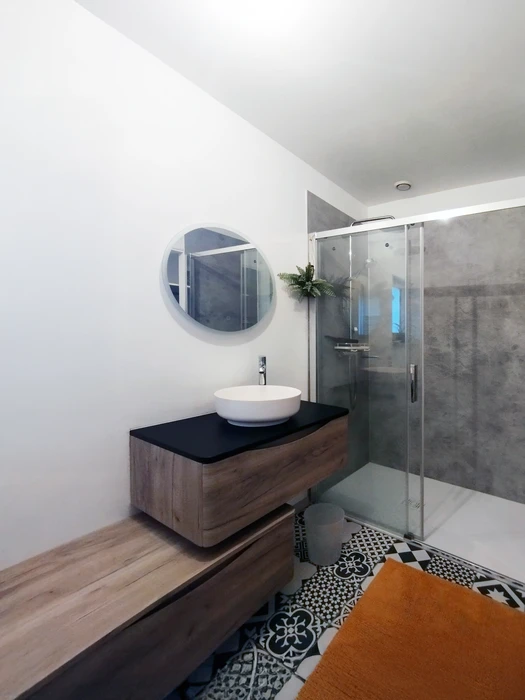 Salle de bain moderne avec douche à l'italienne vitrée, meuble vasque suspendu en bois avec plan noir, vasque blanche ronde, miroir circulal mural