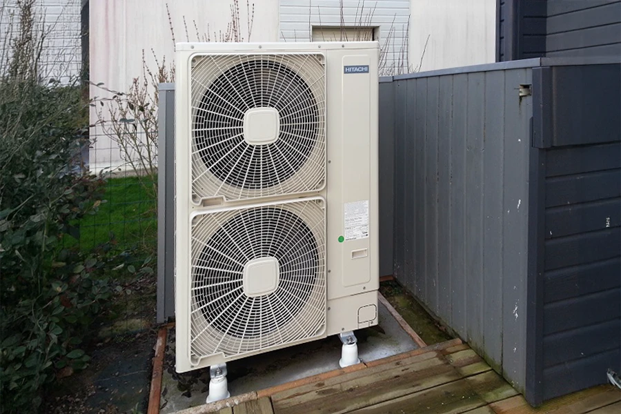 Unité extérieure de pompe à chaleur Hitachi installée sur terrasse en bois, située entre mur béton et clôture métallique grise