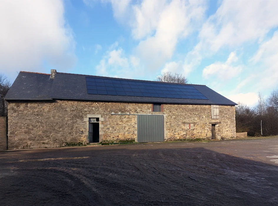 Bâtiment agricole en pierre naturelle avec toiture ardoise équipée de panneaux photovoltaïques, cour bitumée, environnement rural
